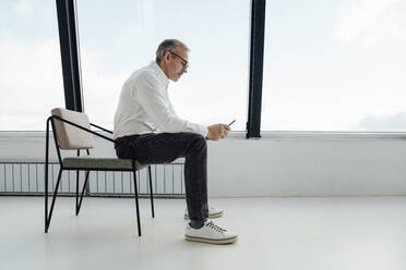 Businessman using smart phone sitting on chair at startup office - VPIF05464