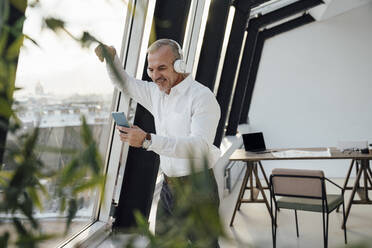 Lächelnder Geschäftsmann, der ein Smartphone am Fenster am Arbeitsplatz benutzt - VPIF05496