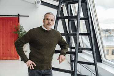 Confident businessman with hand on hip by ladder at work place - VPIF05535