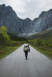 Norway, Lofoten Islands, Backpacker walking down road in mountain landscape - TETF00084