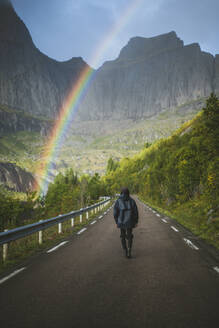 Norwegen, Lofoten-Inseln, Mann geht Straße entlang mit Bergen und Regenbogen im Hintergrund - TETF00091
