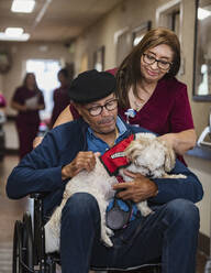 Lächelnde Krankenschwester, die einen älteren Mann mit Hund im Rollstuhl schiebt - TETF00317