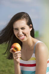 Young woman with apple winking eye and sticking out tongue on sunny day - MIKF00082