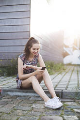 Smiling young woman with smart phone sitting on footpath - MIKF00093