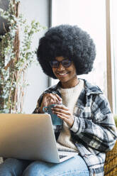 Lächelnde Geschäftsfrau mit Tasse und Laptop auf dem Schoß in einem Café - PNAF03320