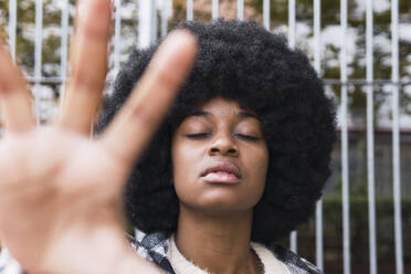 Afro woman with eyes closed showing palm in front of fence - PNAF03349