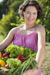 Lächelnde Frau mit Korb mit frischem Gemüse im Park an einem sonnigen Tag - MIKF00103