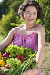 Smiling woman with basket of fresh vegetables in park on sunny day - MIKF00103