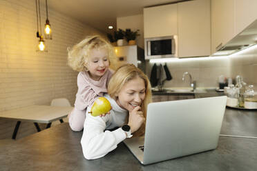 Playful daughter sitting on mother's back in kitchen at home - SIF00049