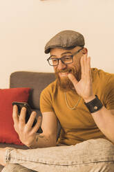 Bearded man having video call on mobile phone sitting in living room at home - MGRF00635