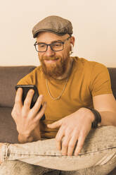 Smiling man holding mobile phone and listening music through in-ear headphones at home - MGRF00636