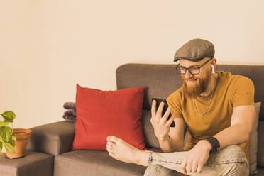 Bärtiger Mann mit Videoanruf auf Smartphone im Wohnzimmer sitzend - MGRF00637