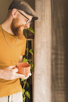 Fröhlicher Mann mit Kaffeetasse am Fenster zu Hause - MGRF00639