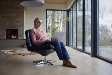 Blonde Frau mit Brille und Laptop auf einem Stuhl im Wohnzimmer sitzend - RBF08539