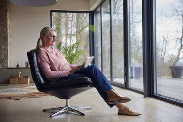 Lächelnde Frau mit Laptop auf einem Stuhl vor einem Glasfenster zu Hause - RBF08540