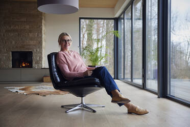 Smiling blond woman with tablet PC sitting on chair at home - RBF08542