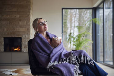 Entspannte, in eine Decke gehüllte Frau sitzt mit geschlossenen Augen im Wohnzimmer - RBF08545