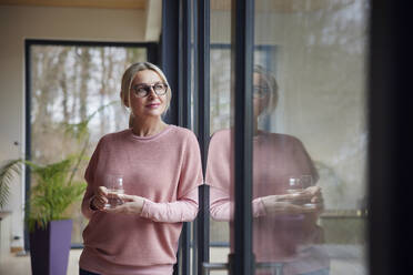 Blonde Frau mit Trinkglas schaut durch ein Fenster zu Hause - RBF08547