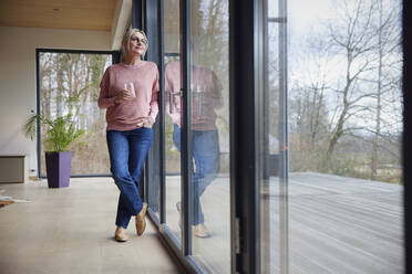 Frau mit Trinkglas und Hand in der Tasche am Fenster stehend - RBF08548