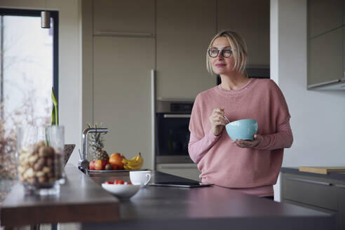 Nachdenkliche blonde Frau mit Schüssel in der Küche zu Hause stehend - RBF08555