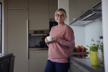 Happy blond woman holding coffee cup standing by kitchen counter at home - RBF08558
