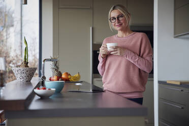 Blond woman wearing eyeglasses holding coffee cup standing in kitchen - RBF08559