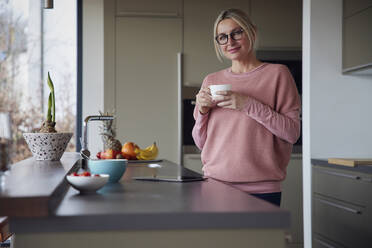 Blonde Frau mit Brille und Kaffeetasse in der Küche stehend - RBF08559