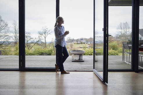 Frau mit Kaffeetasse vor einem Glasfenster zu Hause - RBF08564