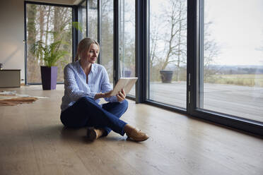 Frau mit Tablet-PC auf dem Boden sitzend vor einem Glasfenster zu Hause - RBF08570