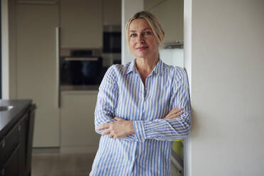 Blond woman standing with arms crossed leaning on wall at home - RBF08572