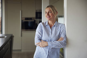 Happy blond woman standing with arms crossed leaning on wall at home - RBF08573