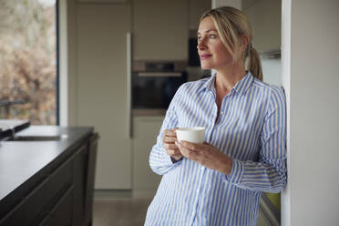 Blond woman holding coffee cup leaning on wall at home - RBF08574