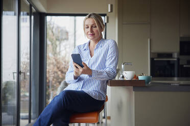 Blonde Frau, die in der Küche sitzend mit ihrem Smartphone im Internet surft - RBF08576