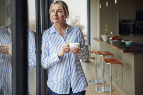 Blonde Frau hält Kaffeetasse und schaut durch das Fenster - RBF08577