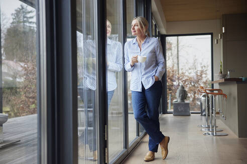 Frau mit Kaffeetasse stehend mit Hand in der Tasche schaut durch das Fenster - RBF08578