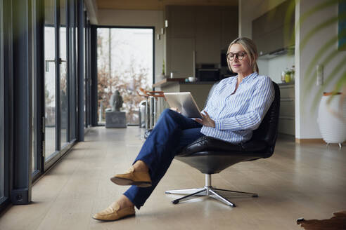 Frau mit Brille und Tablet-PC auf einem Stuhl zu Hause - RBF08581