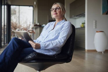 Nachdenkliche Frau mit Brille sitzt mit Tablet-PC auf einem Stuhl zu Hause - RBF08582