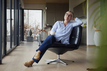 Relaxed woman wearing eyeglasses holding coffee cup sitting on chair at home - RBF08584