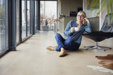 Blonde Frau mit Brille und Kaffeetasse im Schneidersitz auf einem Stuhl zu Hause - RBF08592