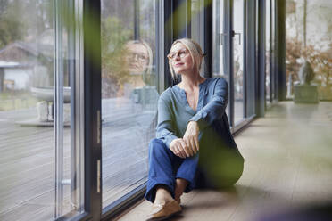 Thoughtful blond woman looking through glass window sitting at home - RBF08593