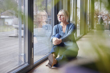 Woman sitting by glass window at home - RBF08594