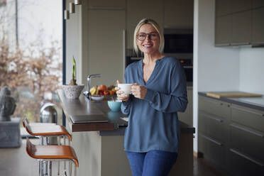 Smiling blond woman holding coffee cup standing in kitchen at home - RBF08598