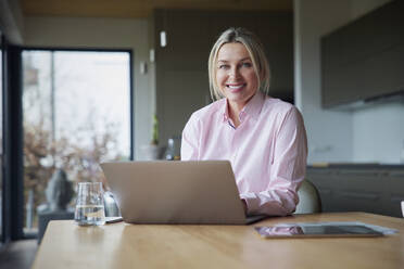 Glückliche blonde Frau sitzt mit Laptop am Tisch - RBF08613