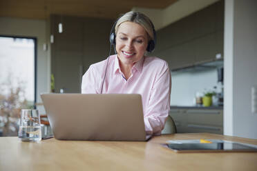 Smiling blond woman wearing headphones using laptop sitting at table - RBF08618