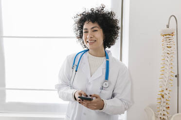 Glücklicher junger Arzt mit Smartphone am Fenster einer Klinik - XLGF02729