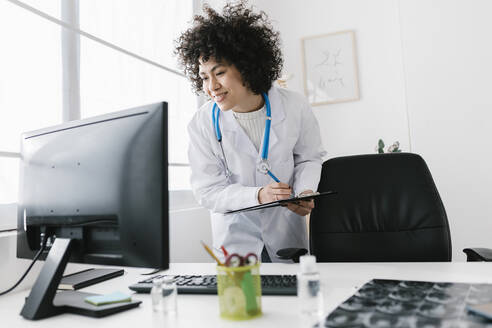 Smiling doctor holding clipboard looking at desktop PC in medical clinic - XLGF02736