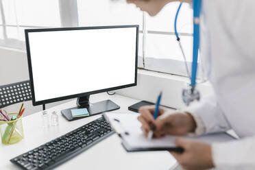 Arzt schreibt auf Klemmbrett und schaut auf den Computerbildschirm in der Klinik - XLGF02739