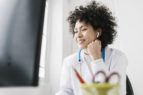 Smiling doctor sitting with hand on chin listening through in-ear headphones at clinic - XLGF02752