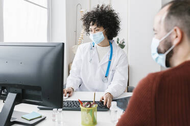 Doctor wearing protective face mask using computer sitting with patient at medical clinic - XLGF02770