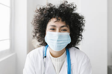 Young doctor with curly hair wearing protective face mask in medical clinic - XLGF02774
